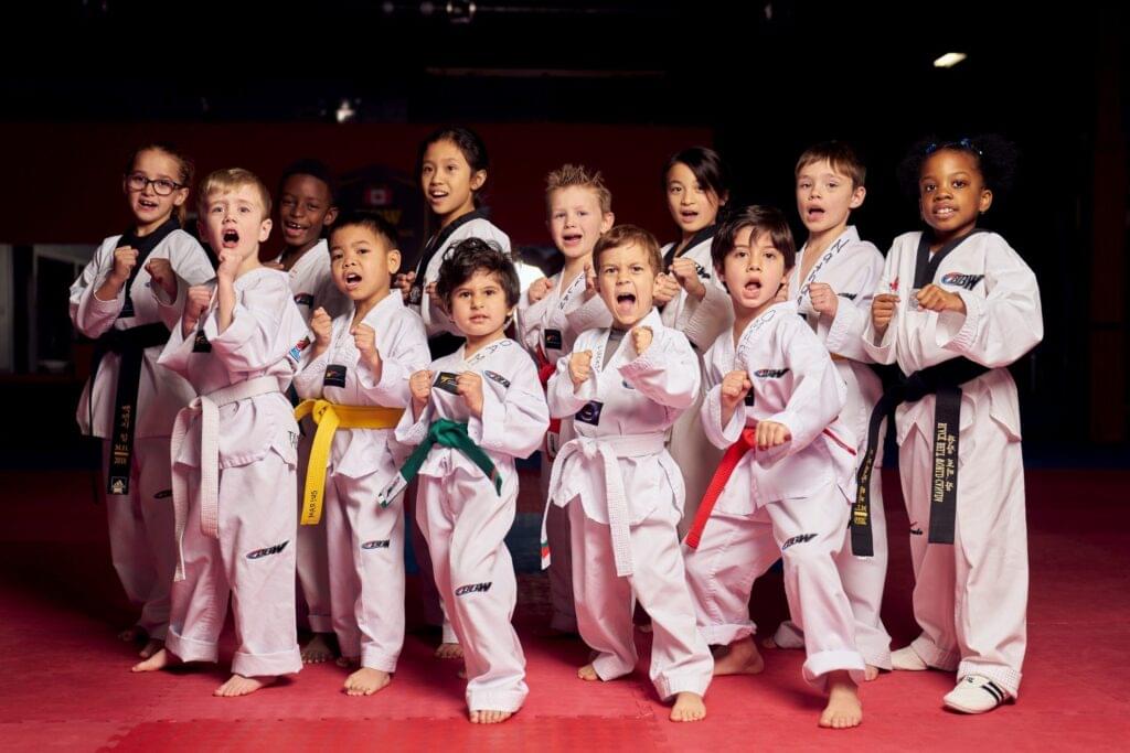 A diverse group of young martial arts students wearing white uniforms and various colored belts practicing their kihap shouts during a kids' karate class.
