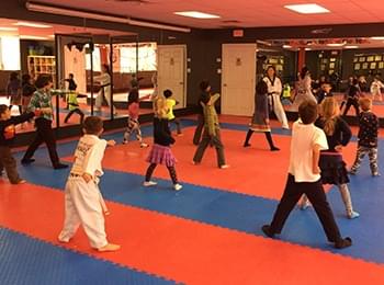 A group of students in a bright taekwondo dojang practicing martial arts forms under the guidance of an instructor.