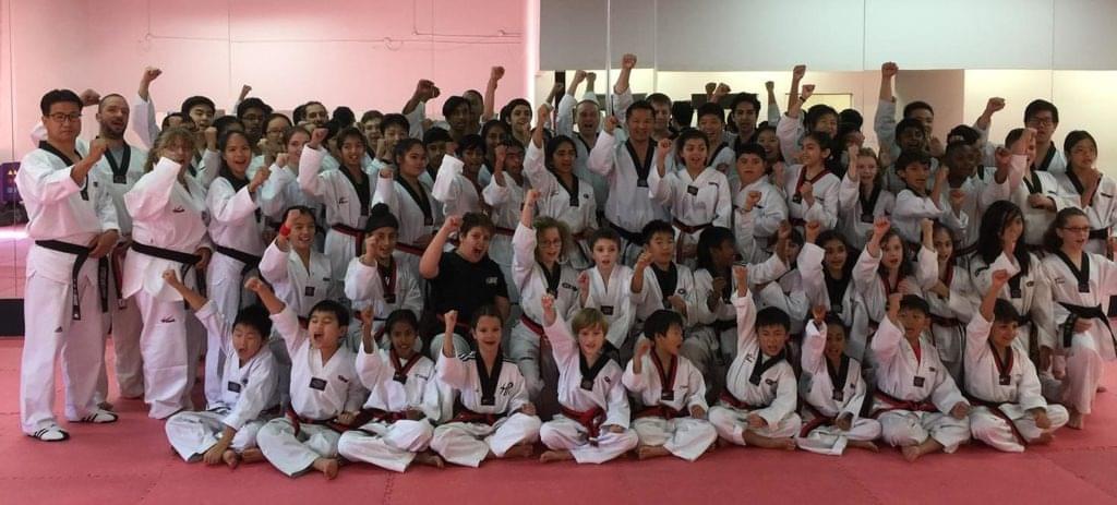 A large, diverse group of students and instructors in white uniforms celebrating with a unified arm raise during a martial arts graduation and community fitness event.